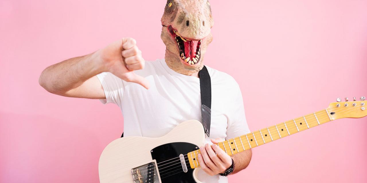 Man with dinosaur mask and guitar around his neck points thumbs down