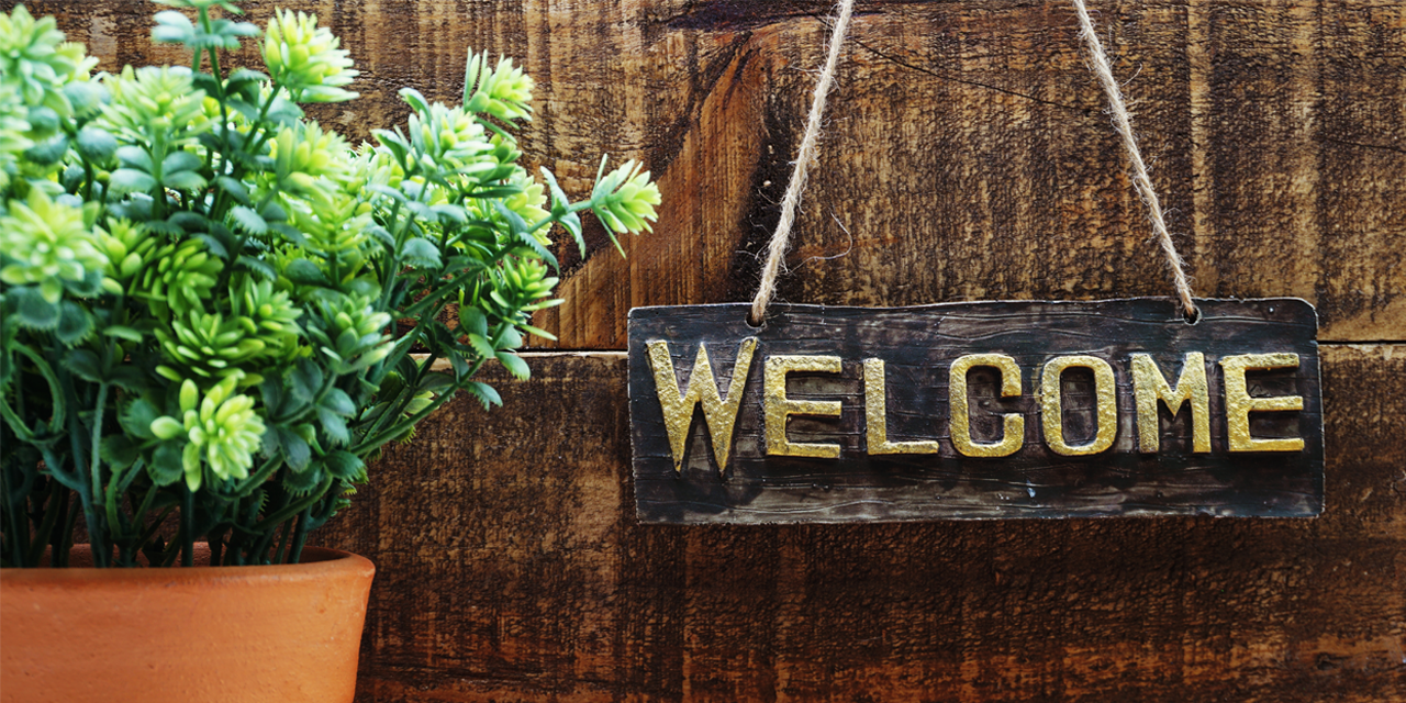 Ein Schild, das auf einem Zaun hängt und auf dem das Wort "Welcome" - Englisch für Willkommen - steht. Daneben steht ein Topf mit einer Pflanze.