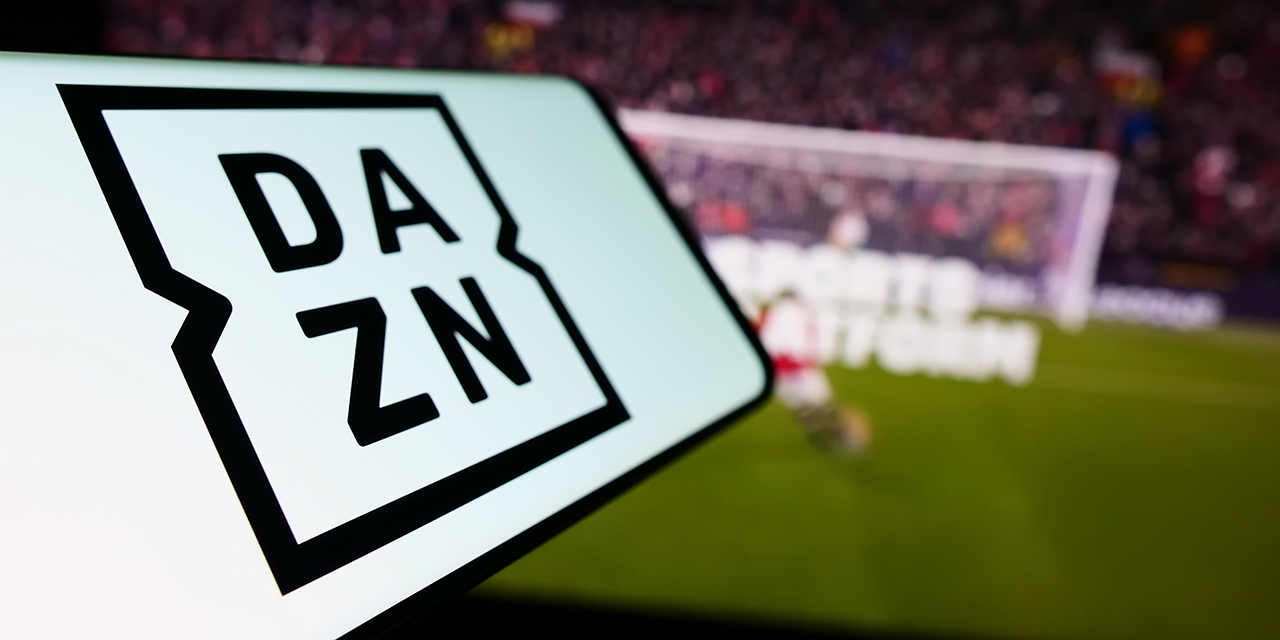 Das Logo von DAZN auf einem Smartphone, dahinter ein Tor im Fußballstadion.