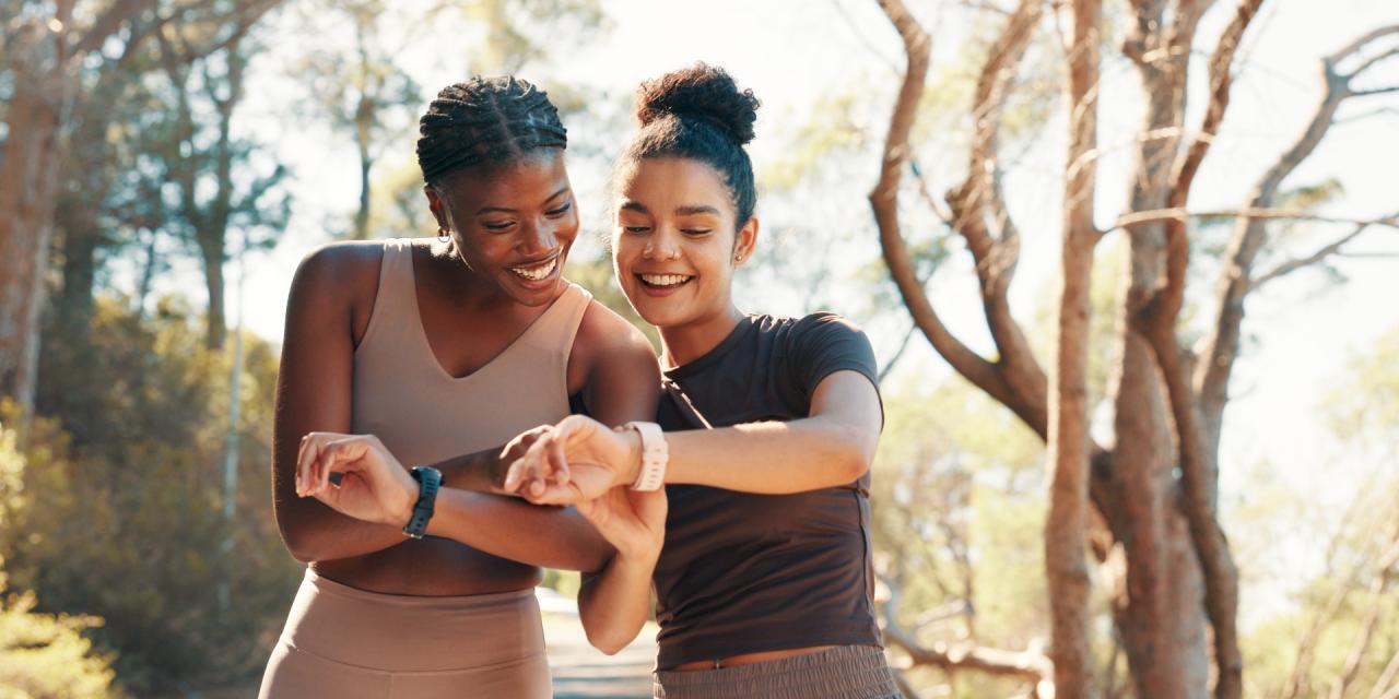 Zwei Frauen in Sportkleidung blicken auf ihre Smartwatches und lachen dabei. 