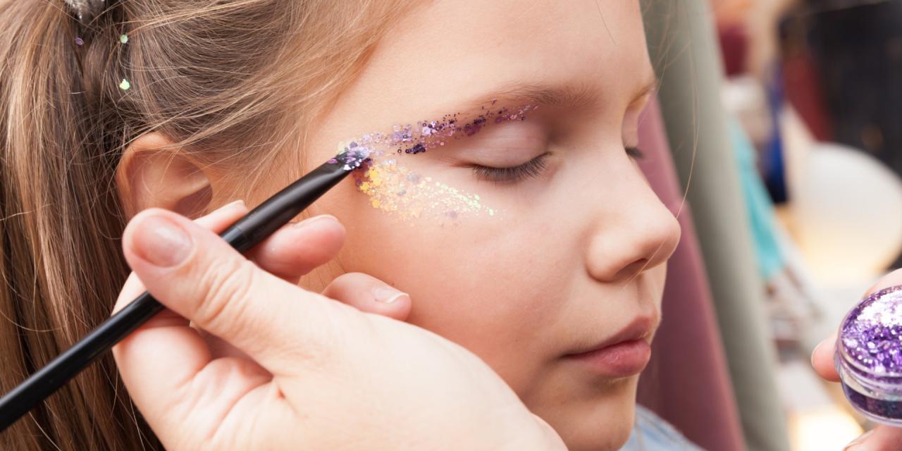 Ein Mädchen wird im Gesicht mit buntem Glitzer geschminkt. 