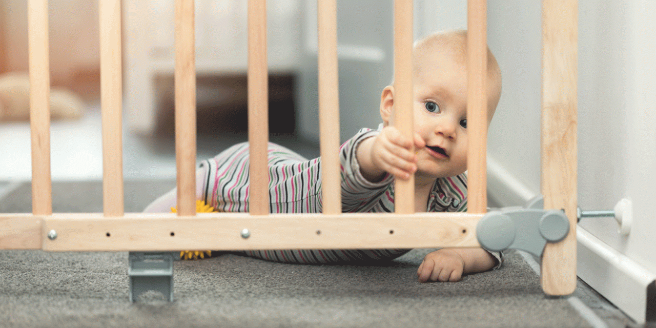 Baby hinter einem Treppenschutzgitter. Das Gitter besteht aus hellem Holz. Das Baby trägt einen gestreiften Einteiler und schaut neugierig durch das Gitter hindurch. Es liegt auf dem Bauch auf einem Teppichboden und greift mit einer Hand nach einer der Holzstreben.