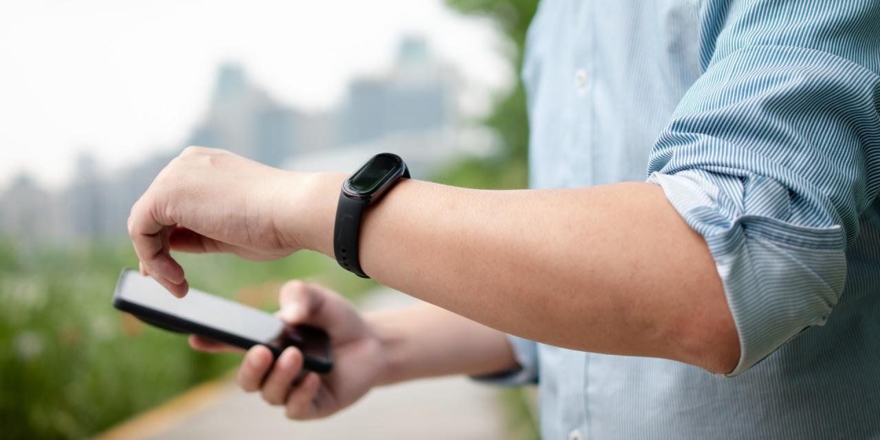 Ein Mann trägt einen Fitnesstracker, in der anderen Hand hält er ein Smartphone, er trägt ein blaues Hemd. 