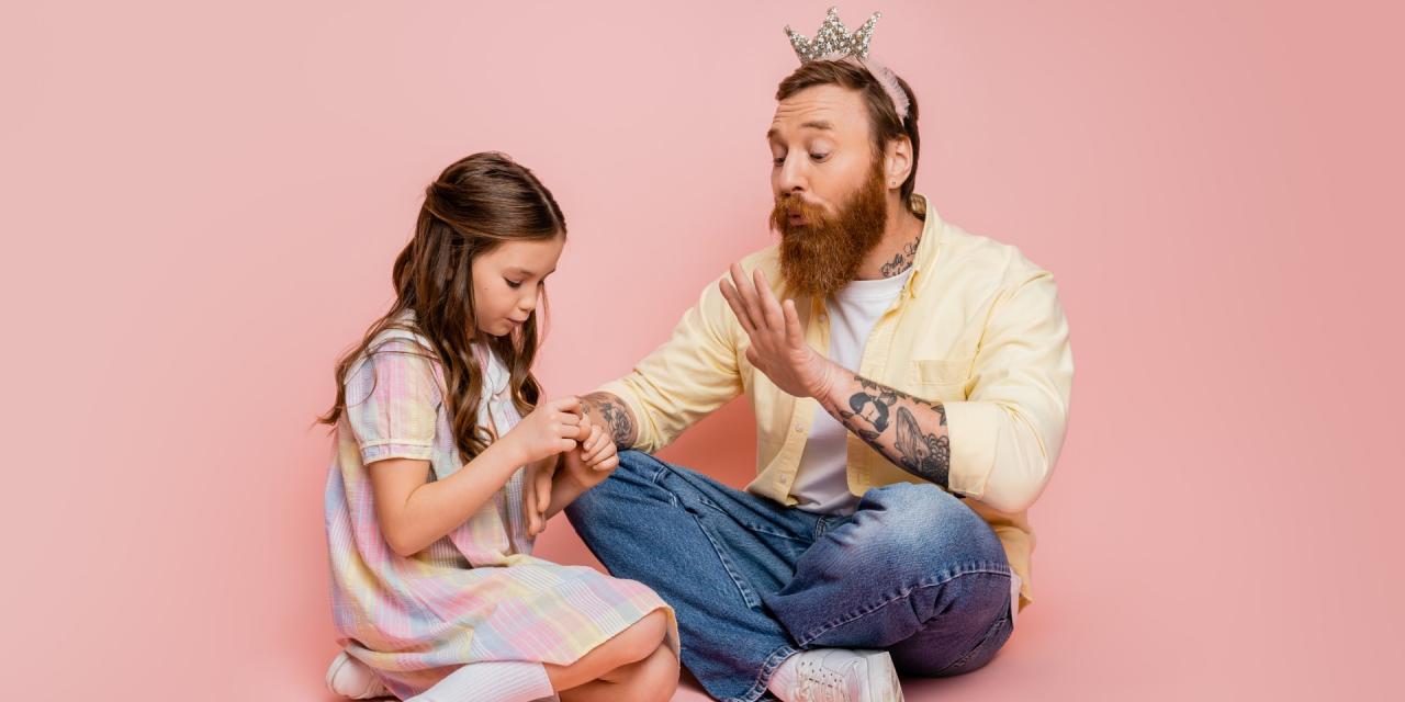 Vater und Tochter sitzen am Boden, der Vater hat eine Krone auf, die Tochter lackiert ihm die Fingernägel