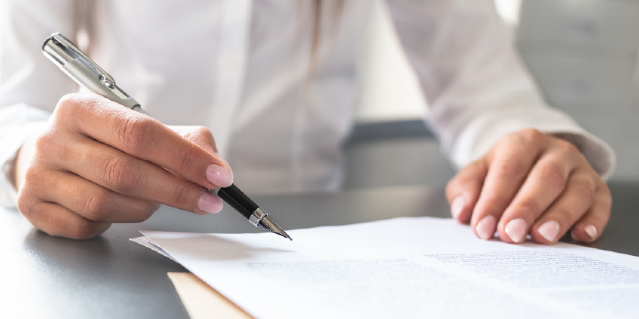 Eine Person hält einen schwarzen Stift in der Hand und schreibt auf ein Blatt Papier, das auf einem Tisch liegt. Der Hintergrund ist unscharf.