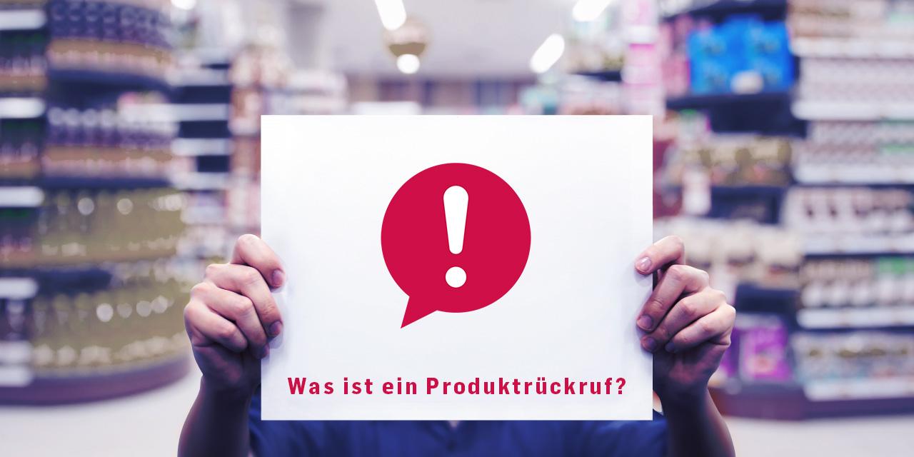 Eine Person hält ein weißes Schild in einem Supermarkt hoch. Auf dem Schild ist ein rotes Symbol mit einem Ausrufezeichen in einer Sprechblase abgebildet. Unter dem Symbol steht der Text: „Was ist ein Produktrückruf?“