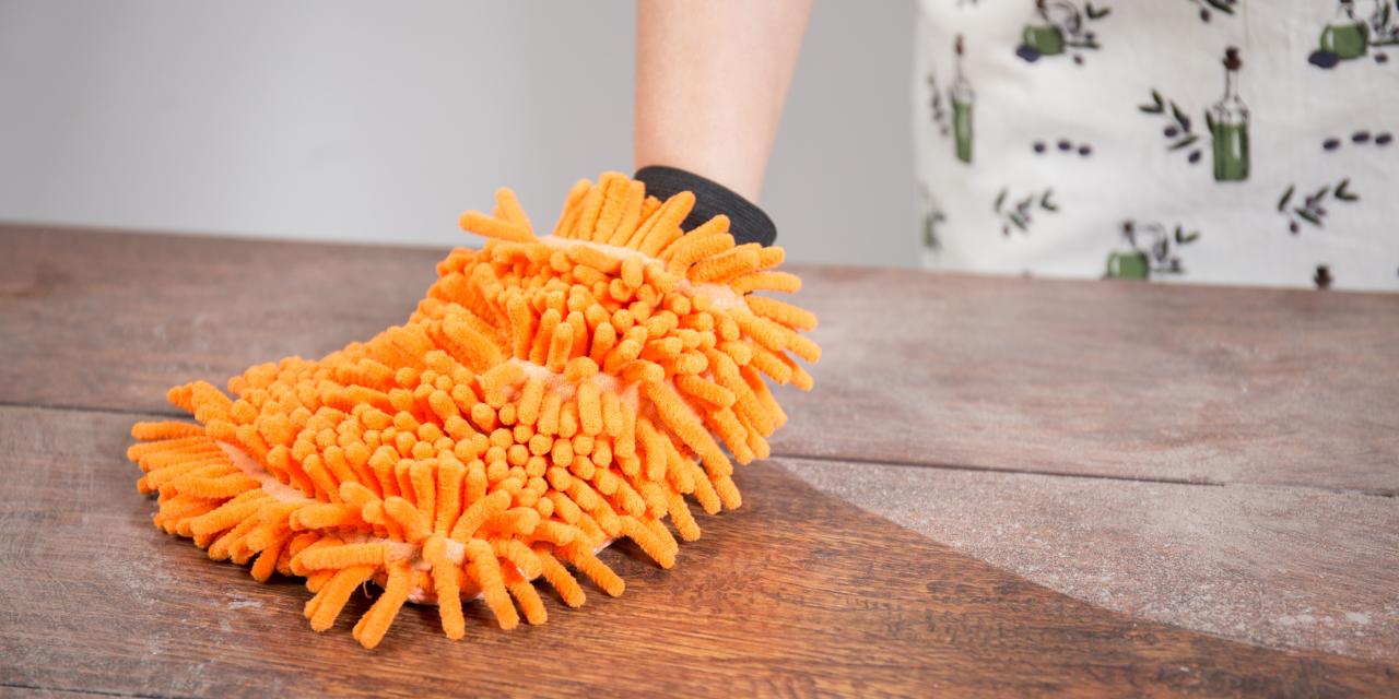 Eine Frauenhand in einem orangen Wischmopp, der wie ein Handschuh anzuziehen ist. Sie wischt damit über einen staubigen Holztisch