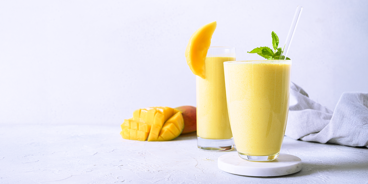 Zwei Gläser mit gelbem Mango-Trinkjoghurt stehen auf einer hellen Oberfläche. Ein Glas ist mit einem Stück Mango garniert, das andere mit frischer Minze und einem Strohhalm. Daneben liegt eine in Würfel geschnittene Mango.