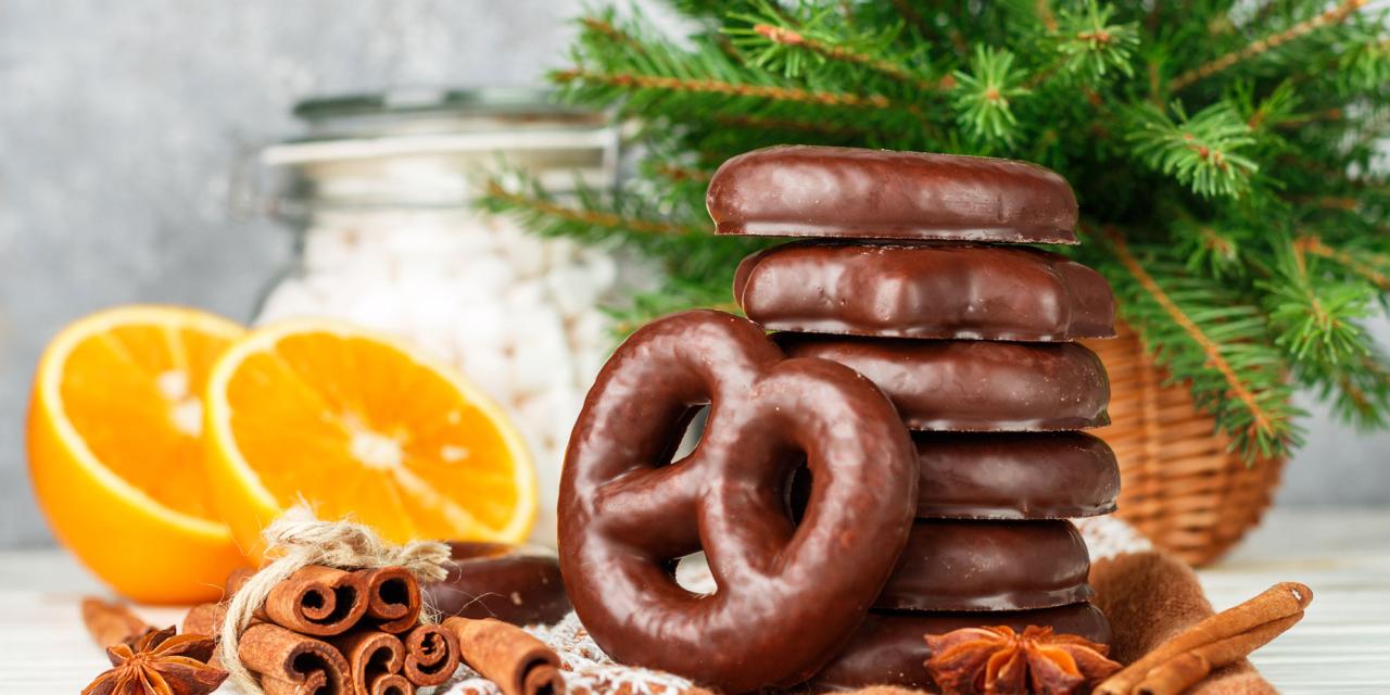Schokoladeüberzogene Lebkuchen gestapelt und aufgestellt vor Christbaumzweigen und 2 Orangenhälften, neben Zimtstangen