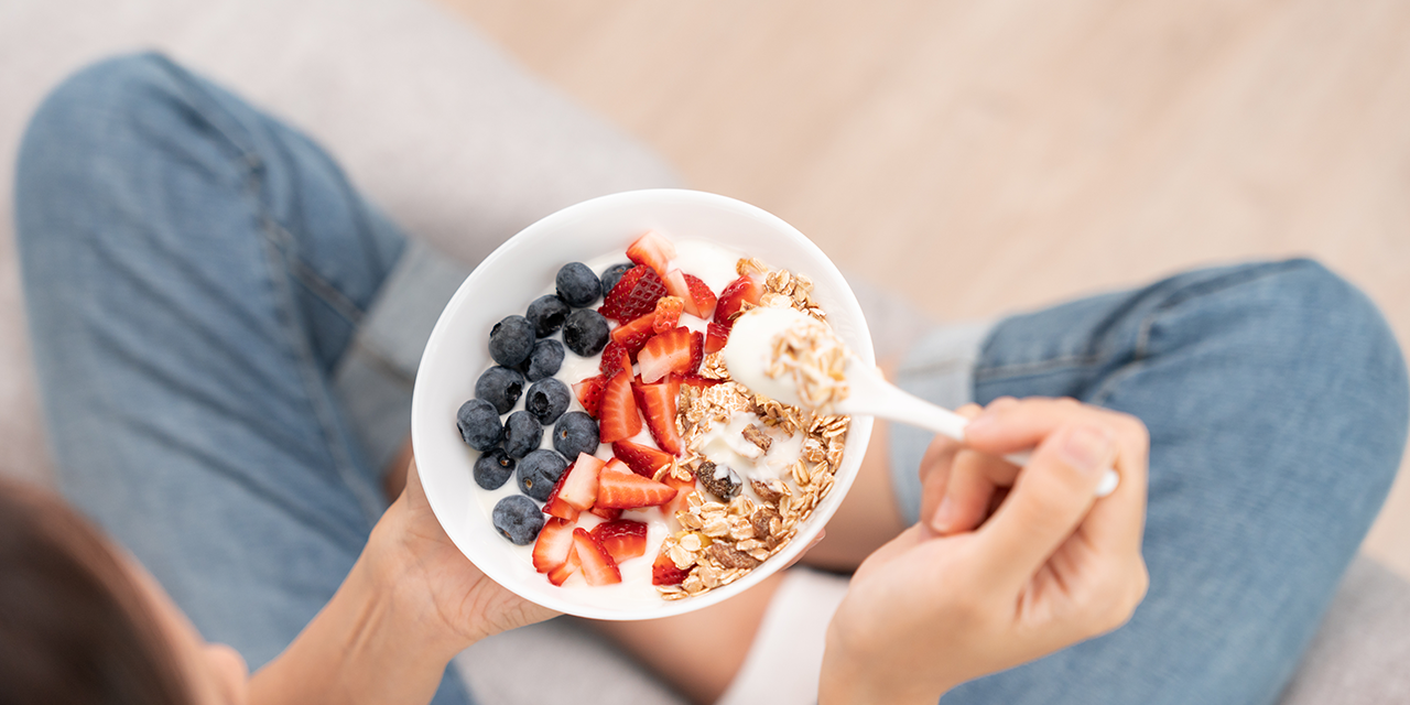 Eine Person hält eine Schüssel mit Naturjoghurt, frischen Heidelbeeren, geschnittenen Erdbeeren und Müsli in den Händen. Ein Löffel mit Joghurt und Müsli wird gerade zum Mund geführt.