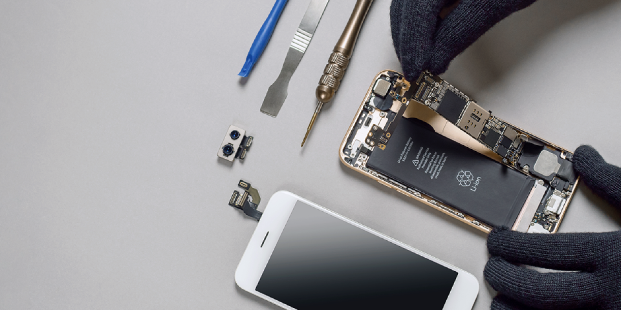 Eine Person mit schwarzen Handschuhen öffnet ein Smartphone und arbeitet am Inneren des Geräts. Rundherum liegen Werkzeug, Ersatzteile und ein zweites Handy auf einer grauen Oberfläche.
