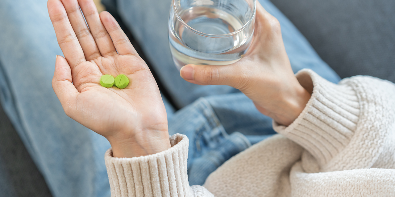 Person hält zwei grüne Tabletten in der einen Hand und ein Glas Wasser in der anderen. Die Person trägt einen hellen Strickpullover und sitzt entspannt auf dem Sofa.
