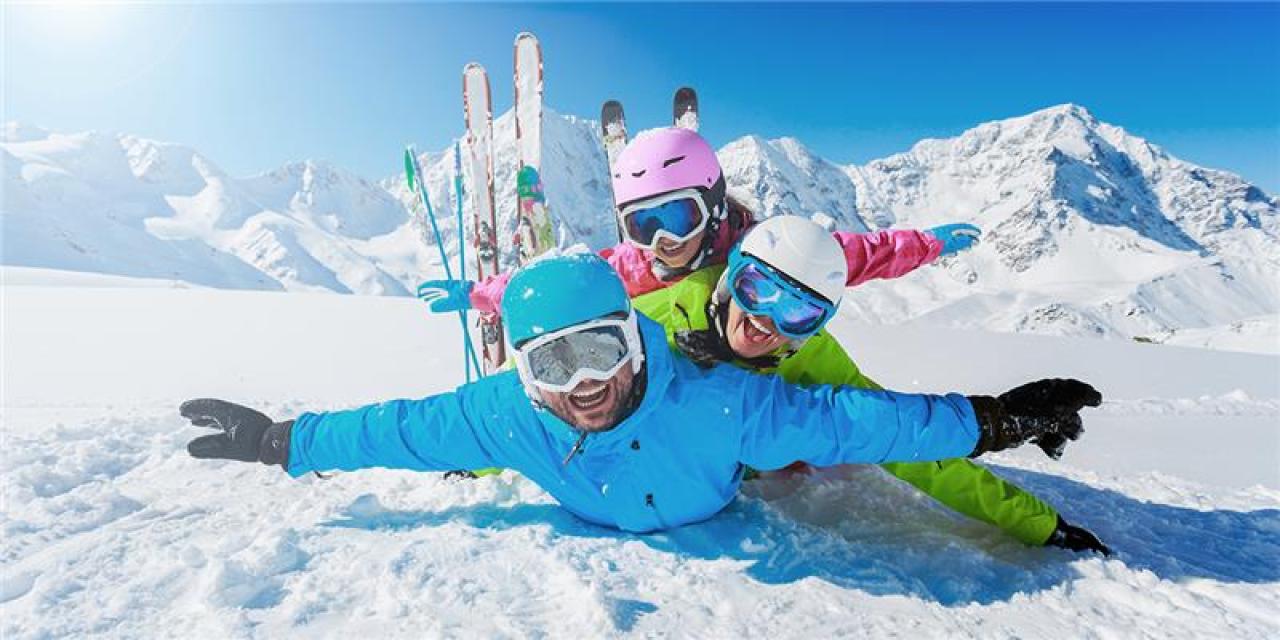 Drei fröhliche Menschen in bunter Ski-Ausrüstung liegen übereinander im Schnee und breiten spielerisch die Arme aus; im Hintergrund stehen Skier im Schnee und schneebedeckte Berge leuchten in der Sonne.