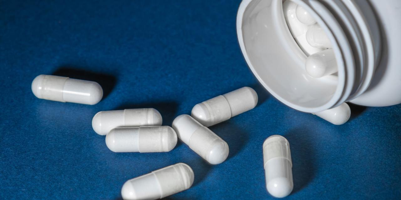 Blauer Untergrund, darauf sieben verstreute weiße Kapseln einer Nahrungsergänzung und angeschnitten eine offene, liegende Plastikdose, in der weitere Kapseln sichtbar sind