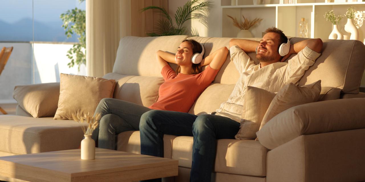 Zwei Personen sitzen entspannt auf einem beigen Ecksofa in einem hellen Wohnzimmer. Beide tragen weiße Over-Ear-Kopfhörer und lehnen sich mit hinter dem Kopf verschränkten Armen zurück. Der Raum ist in neutralen Tönen gehalten, mit Sonnenlicht von links, einem Holztisch mit Vase und einem Regal mit Büchern, Pflanzen und Deko.