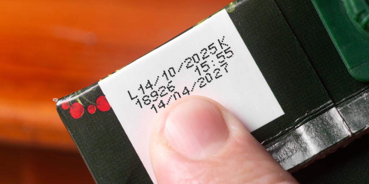 Ein Finger zeigt auf ein weißes Etikett auf einer dunkelgrünen Verpackung. Auf dem Etikett sind mehrere in Schwarz gedruckte Zahlen- und Datumsangaben zu sehen, darunter Produktions- oder Haltbarkeitsdaten. Der Hintergrund ist leicht unscharf, sodass der Fokus klar auf dem Etikett und dem Finger liegt. Die Verpackung zeigt am Rand ein kleines dekoratives Muster.