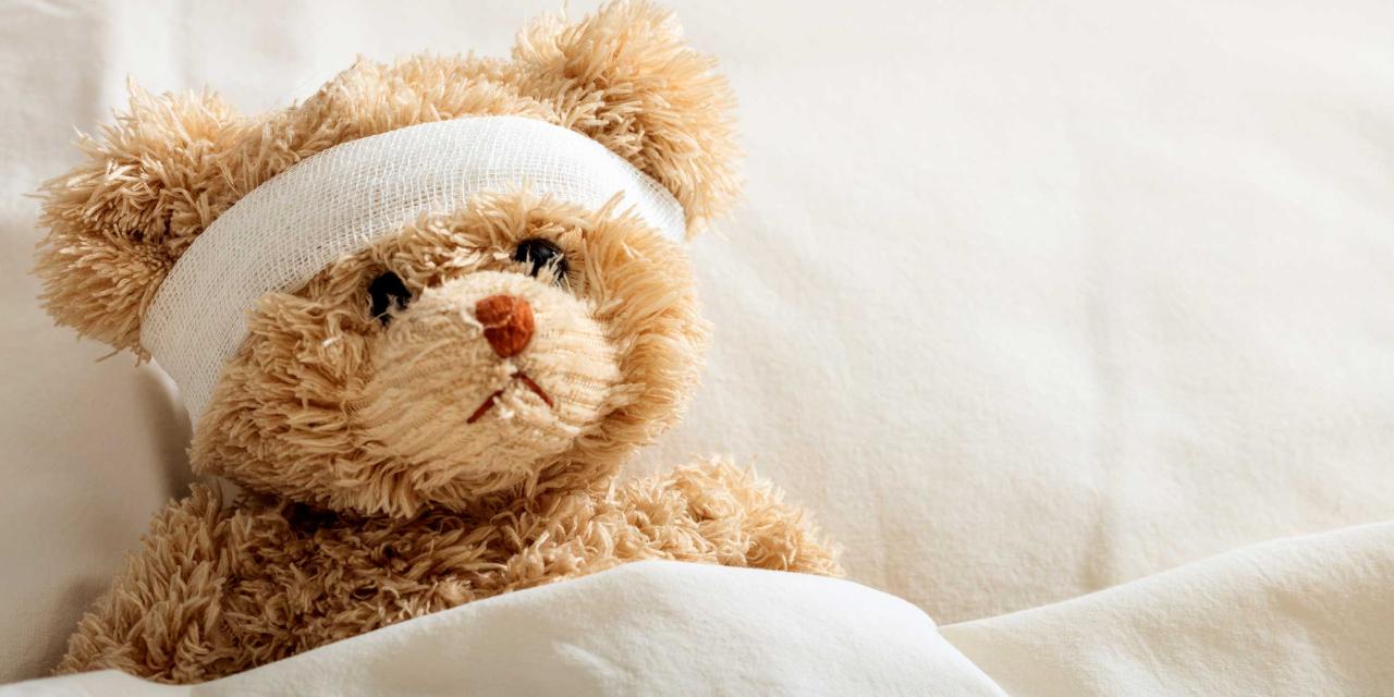 Teddybär liegt mit verbundenem Kopf in einem Spitalsbett