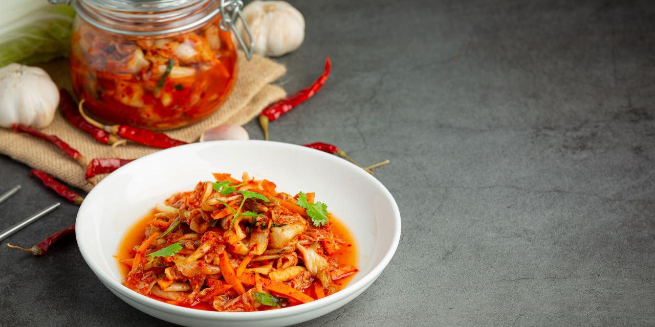 Ein Teller mit Kimchi, dahinter ein Glas gefüllt mit Kimchi, daneben Knoblauch; alles steht auf einer grau melierten Arbeitsplatte