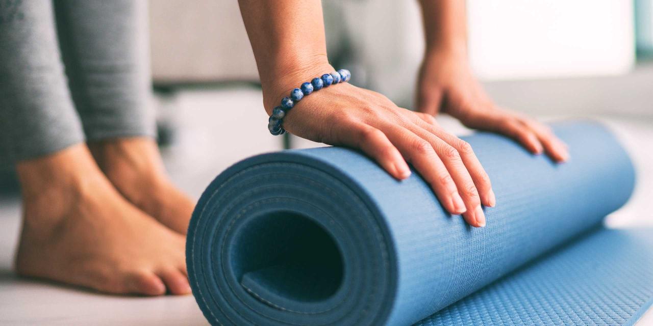 Frau mit Leggings und nackten Füßen rollt blaue Yogamatte am Boden aus