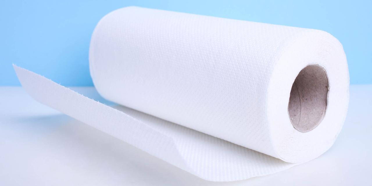 Das Bild zeigt eine weiße Küchenpapierrolle auf einer glatten, hellen Oberfläche vor blauem Hintergrund. Ein Stück des Papiers ist von der Rolle abgerollt und liegt locker nach vorne ausgebreitet. Die Struktur des Papiers wirkt weich und leicht geprägt.