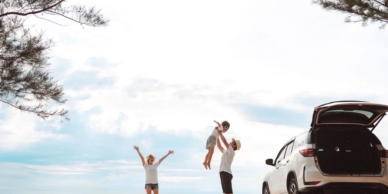Auto am Strand geparkt, Kofferraum geöffnet, Elternteil reißt Arme in die Höhe, anderer Elternteil schwingt Kind in der Luft, Wolken, Meer im Hintergrund