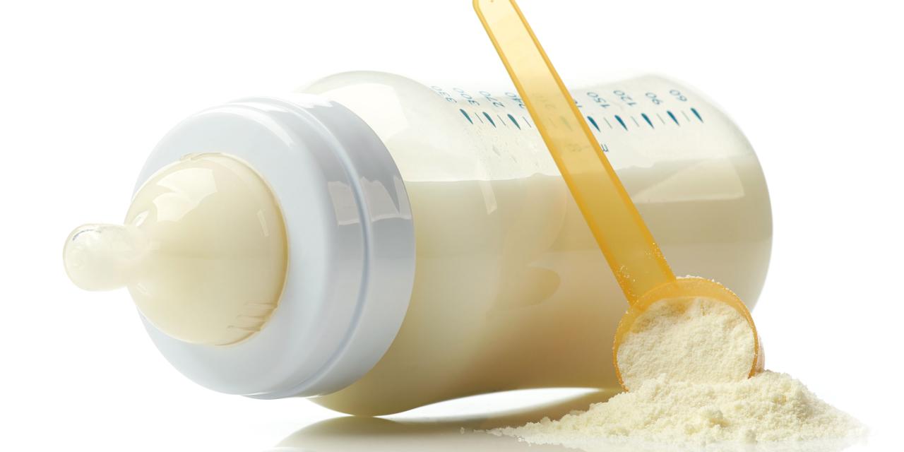 Eine gefüllte Babytrinkflasche liegt auf der Seite. Eine Portionslöffel voll mit pulverförmiger Säuglingsnahrung ist daran angelehnt.