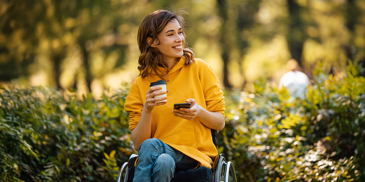 Eine junge Frau im Rollstuhl ist in einem Wald. Sie hält einen Kaffeebecher und ein Smartphone in den Händen.