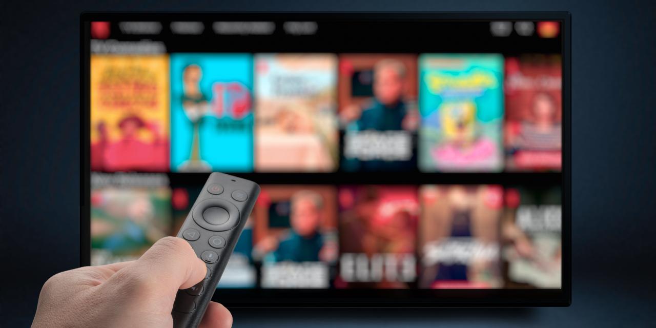 Eine Person hält eine Fernbedienung in der Hand, die sie auf einen verschwommen im Hintergrund befindlichen Fernseher richtet.