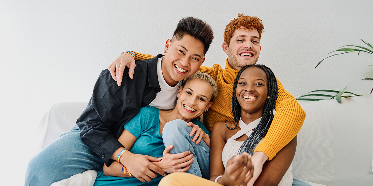 Vier junge Erwachsene sitzen eng zusammen auf einem Sofa und lächeln in die Kamera. Die Gruppe wirkt vertraut und entspannt; im Hintergrund ist eine helle Wand mit einer Pflanze zu sehen.