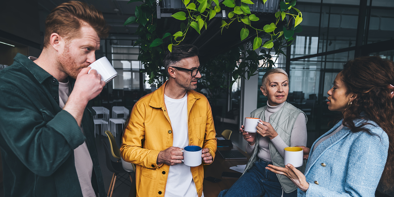 Vier Erwachsene stehen in einem Büro zusammen, halten Tassen und unterhalten sich. Im Hintergrund sind Pflanzen und ein moderner Arbeitsbereich zu sehen.