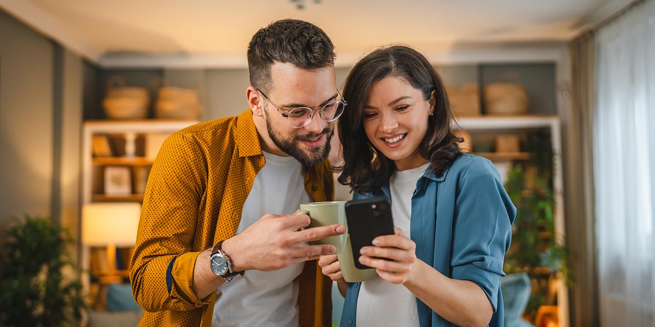 Ein paar steht gemeinsam im Wohnzimmer und sie sehen sich zusammen etwas am Smartphone an.