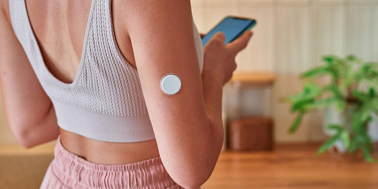 Rücken einer Frau mit Sportoutfit, am Oberarm ein Glucosemessgerät, Handy in der Hand, Küchenanrichte im Hintergrund 