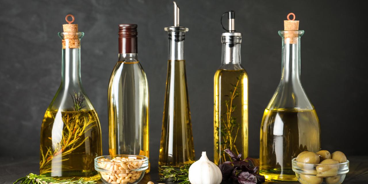 5 neutrale Glasflaschen, befüllt mit verschiedenen Ölen und Kräutern. Davor liegen Nüsse, Knoblauch und Oliven auf dem Tisch