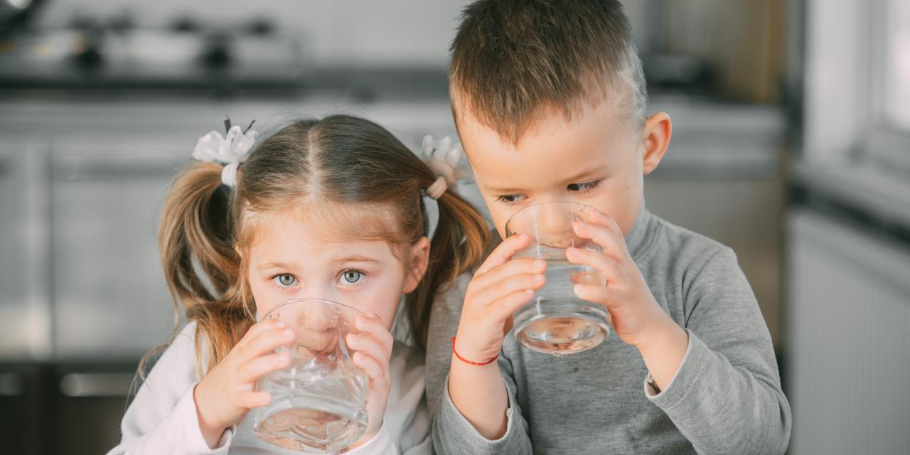 Mädchen und Junge trinken aus Wasserglas beim Holztisch, Küche im Hintergrund verschwommen