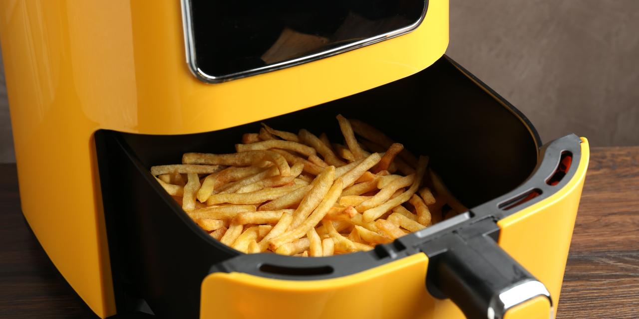 Gelbe Heißluftfritteuse mit herausgezogenem Korb auf einem Holztisch, darin goldbraun frittierte Pommes frites. Über dem Korb ein schwarzes Sichtfenster.