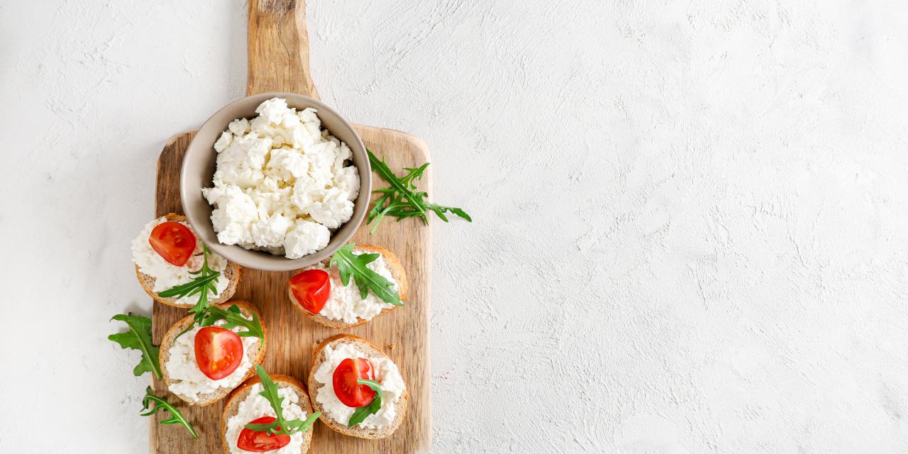 Holzbrett mit Schale Frischkäse und kleinen Brotscheiben, belegt mit Frischkäse, Tomaten und Rucola, auf hellem Hintergrund.