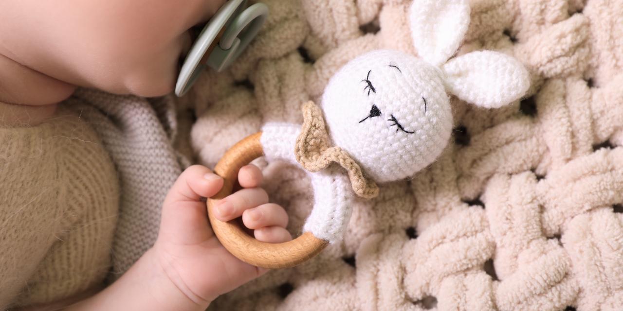 Baby mit Schnuller hält eine gehäkelte Hasenrassel mit Holzring und liegt auf einer weichen, hellen Strickdecke.