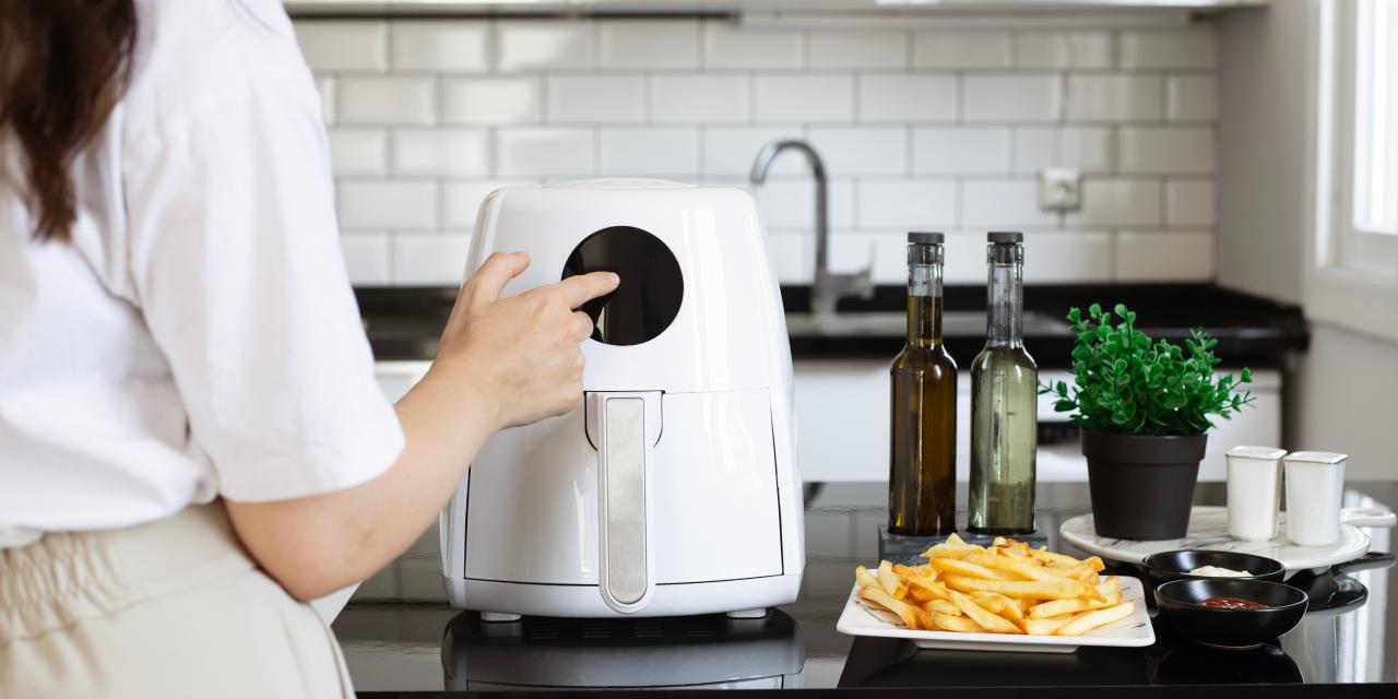 Eine Frau bedient in einer Küche eine weiße Heißluftfritteuse. Daneben ein Teller Pommes frites mit Ketchup und Mayonnaise sowie zwei Ölflaschen.