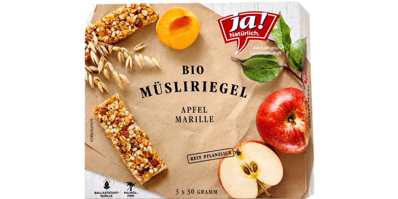 Faltschachtel des „ja! Natürlich" Bio-Müsliriegels „Apfel Marille" zu 3 x 30 Gramm in Schrägansicht, daneben Marille, Apfel und Hafer, mit den Hinweisen „rein pflanzlich" und „palmölfrei".