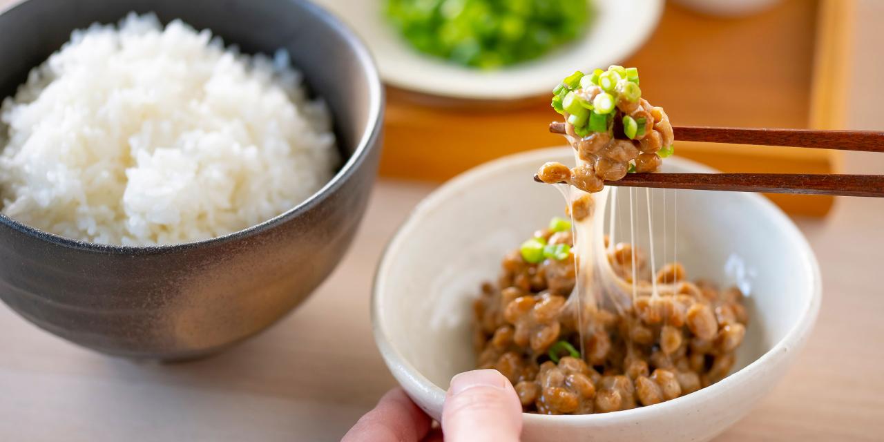 Graue Schüsssel mit Reis, daneben hält eine Hand eine weiße Schüssel mit Natto und Stäbchen entnehmen einen Bissen, Im Hintergrund Tablett mit weißem Teller, der grüne Speise enthält.