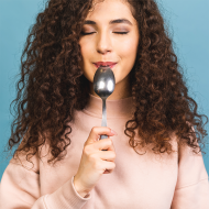 Eine Frau mit lockigem Haar hält einen Löffel vor ihren Mund und lächelt mit geschlossenen Augen. Sie trägt ein helles Oberteil; der Hintergrund ist einfarbig blau.