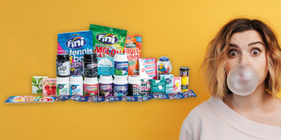 Frau mit Kaugummiblase vor dem Mund vor gelbem Hintergrund, links im Bild Kaugummipackungen.