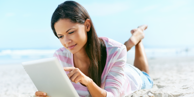 Eine Frau mit einem Tablet in der Hand, die am Strand liegt.