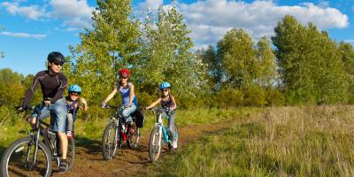 TEST Kinderfahrradhelme: 1 Erwachsener Radfahrer mit Kind am Fahrradsitz, 1 Kind am Kinderfahrrad, 1 weitere Erwachsene am Fahrrad, alle tragen Fahrradhelme