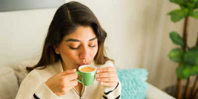 Auf Sofa sitzende dunkelhaarige junge Frau spitzt die Lippen an der gefüllten Espressotasse in ihren Händen