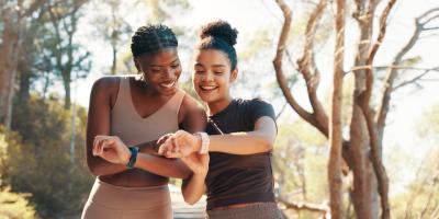 Zwei Frauen in Sportkleidung blicken auf ihre Smartwatches und lachen dabei.