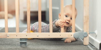 Baby hinter einem Treppenschutzgitter. Das Gitter besteht aus hellem Holz. Das Baby trägt einen gestreiften Einteiler und schaut neugierig durch das Gitter hindurch. Es liegt auf dem Bauch auf einem Teppichboden und greift mit einer Hand nach einer der Holzstreben.