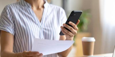 Eine jüngere Frau, deren Gesicht nicht zu sehen ist, hält in der einen Hand ein Handy und in der anderen eine Papierrechnung.
