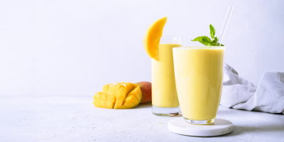 Zwei Gläser mit gelbem Mango-Trinkjoghurt stehen auf einer hellen Oberfläche. Ein Glas ist mit einem Stück Mango garniert, das andere mit frischer Minze und einem Strohhalm. Daneben liegt eine in Würfel geschnittene Mango.