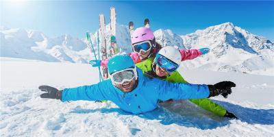 Drei fröhliche Menschen in bunter Ski-Ausrüstung liegen übereinander im Schnee und breiten spielerisch die Arme aus; im Hintergrund stehen Skier im Schnee und schneebedeckte Berge leuchten in der Sonne.